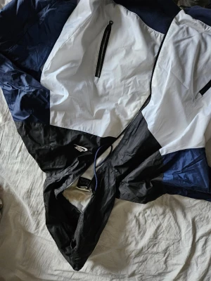 Nike vindjacka i svart, vit och blå - Nike vindjacka i färgblock med svart, vit och marinblå. Jackan har huva, hel dragkedja och två fickor med dragkedja framtill. Lätt och sportig design i syntetmaterial, perfekt för blåsiga dagar. Snygg logga på bröstet och meshfoder på insidan.