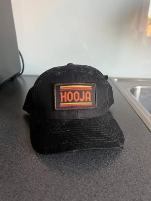 Svart Hooja keps med justerbar rem - Snygg svart keps från Hooja med broderad patch framtill i orange och gult. Kepsen har klassisk form, böjd skärm och justerbar plastrem baktill. Insidan har Hooja-logga på bandet och etikett. Perfekt för dig som vill ha en cool och enkel accessoar.