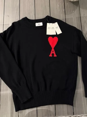 Svart tröja med rött hjärta från Ami - Snygg svart tröja från Ami med ett stort rött hjärta och bokstaven A broderat på bröstet. Tröjan har rund hals, ribbade muddar och är långärmad. Perfekt för dig som gillar stilrena plagg med en unik detalj.