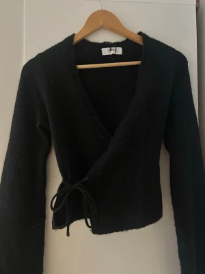 Svart omlottkofta från Gina Tricot - Svart omlottkofta från Gina Tricot i mjuk ullblandning. Koftan har lång ärm, v-ringad hals och knytband i midjan för en snygg passform. 