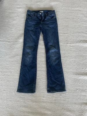 Blå bootcut jeans med fickdetaljer - Säljer ett par mörkblå bootcut jeans med snygga fickdetaljer bak och klassisk femficksdesign. En knapp på ena fickan har gått av. Jeansen har normal passform och är tillverkade i mjukt denimtyg. Perfekta till sneakers eller boots för en avslappnad look.