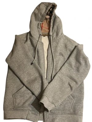 Grå Burberry hoodie med dragkedja - Snygg grå hoodie från Burberry med klassiskt rutigt foder i huvan och insidan. Tröjan har dragkedja, snörning i huvan och broderad Burberry-logga på bröstet. Tillverkad i mjuk bomull med elastan för extra komfort. Perfekt för chill dagar och streetstyle.
