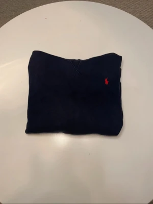 Blå Polo Ralph lauren hoodie  - Mörkblå hoodie från Polo Ralph Lauren i klassisk och tidlös design. Tröjan har huva, känguruficka framtill och den ikoniska röda Polo-loggan broderad på bröstet. Tillverkad i mjukt och bekvämt material med borstad insida som ger extra värme och komfort. Normal passform som passar perfekt till vardagsbruk. Storleken är XL/TG men passar till en kille som är 160 cm lång.