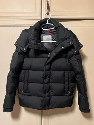 Svart Moncler Vezere Jacka - Svart pufferjacka från Moncler med huva och quiltad design. Jackan har dragkedja och knappar framtill, samt två fickor med dragkedja. Perfekt för kalla dagar och har en stilren look som funkar till många outfits. Storlek 1/S, använd en gång men den är helt felfri och fläckfri. Fraktar snabbt så den hinner komma innan jul. NFC scannar.