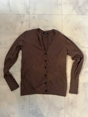 Brun kofta från Gina Tricot - Mysig brun kofta från Gina Tricot i storlek S. Koftan har v-ringad hals, långa ärmar och knappar hela vägen framtill samt extra knappar vid ärmsluten. Perfekt för lager på lager under kyliga dagar.