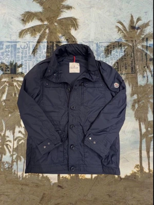 Mörkblå vindjacka från Moncler - Snygg mörkblå vindjacka från Moncler med klassisk logga på ärmen. Jackan har flera fickor, knappar och dragkedja framtill samt hög krage. Materialet är polyester och jackan har en normal passform med långa ärmar. Perfekt för dig som gillar stilrena och funktionella plagg. Storlek 1. Sitter som S/M