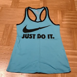 Nike blå linnetopp med svart tryck - Nike Dri-Fit linnetopp i ljusblått med svarta kanter och stort svart Swoosh-tryck samt texten 'JUST DO IT.' framtill. Sportig racerback-modell som är perfekt för träning. Materialet är mjukt och andas, vilket gör den skön att ha på sig under aktiva dagar.