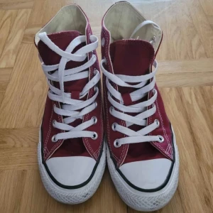 Converse Chuck Taylor vinröda high tops - Säljer ett par klassiska Converse Chuck Taylor All Star high tops i vinrött med vit gummisula och vita snören. Skorna har rund tå, tydliga vita sömmar och det ikoniska All Star-märket på sidan. Materialet är slitstark bomullscanvas och sulan är platt.
