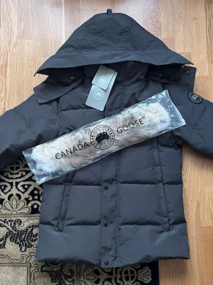 Canada Goose Wyndham Parka – Grå  - Säljer en grå Canada Goose Wyndham Parka i storlek M med svart badge. Jackan är ny, aldrig använd och tags sitter kvar. Det är en varm och stilren vinterjacka med huva och avtagbar päls, perfekt för kalla vinterdagar både till vardag och stadsmiljö. Passar även perfekt som julklapp till kompis eller till era barn. Säljs då den inte kommer till användning.