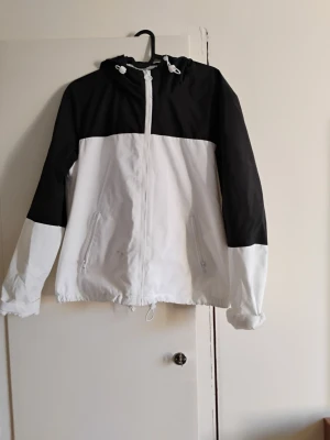 Svartvit vindjacka från FB Sister - Snygg svartvit vindjacka från FB Sister i storlek S. Jackan har huva med snörning, dragkedja framtill och två fickor med dragkedja. Färgblockad design i svart och vitt, samt justerbar nederkant. Perfekt för dig som gillar en sportig och clean stil.jackan har en liten svart fläck därav priset.