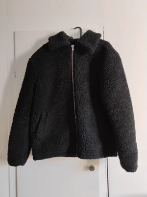 Svart teddyjacka från T.T i S - Mjuk och fluffig svart teddyjacka från T.T i storlek S. Jackan har en klassisk krage, dragkedja framtill och två sidofickor. Perfekt för dig som gillar chill och comfy stil. Materialet är fleece och jackan är långärmad med loose passform.