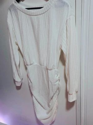 Ribbad Vit Klänning Draperad midja Bodycon M  - Säljer en oanvänd, figurnära klänning i mycket fint skick.  Klänningen är i en härlig, stretchig och ribbad kvalitet i färgen vit/off-white. Den har en smickrande design med en bred/båtformad halsringning, lång ärm och en insydd resår i midjan. Klänningens snyggaste detalj är den draperade/rynkade (ruching) sidan som ger en assymetrisk nederkant och en vacker, figurnära silhuett (bodycon). Perfekt som festklänning, till middag eller som en längre tunika.  Skick: Mycket fint skick, oanvänd. 