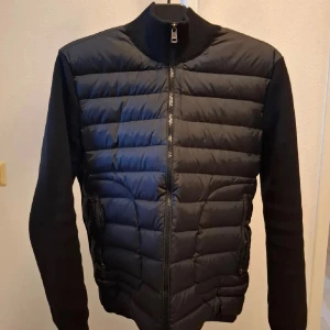 Svart Moncler cardigan jacka - Svart cardigan jacka från Moncler med quiltad front och stickade ärmar. Jackan har dragkedja framtill, ribbad krage och två fickor med dragkedja. Den är använd ett fåtal gånger och är i en mycket fin skick. Passar M/L skulle säga passar perfekt till M, Kvitto finns vid behov!!