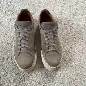 Grå mockasneakers från Strömberg - Snygga grå sneakers från Strömberg i mocka med vita sulor och klassisk snörning. Skorna har en stilren design med rund tå och diskret branding på plösen. Perfekta för dig som gillar en clean och enkel look.