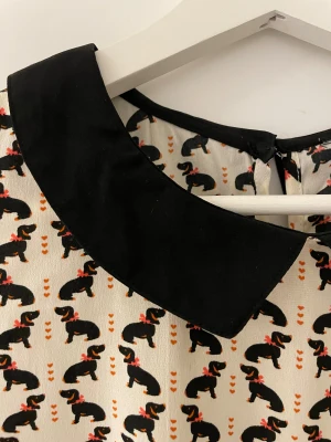 Ärmlös klänning med taxmönster  - Säljer en söt ärmlös klänning med svart krage och ett unikt mönster med taxar och små hjärtan. Basen är vit med svarta detaljer och taxarna har röda rosetter.                                                Klänningen har en rundad krage och lätt utställd kjol. Perfekt för dig som gillar djurmotiv och vill sticka ut. Klänningen har ingen stl men jag uppskattar den till stl 44. Se mått.  Bröst: 58 cm   Midja: 57 cm Längd: 90 cm Armhåla: 30 cm                                 