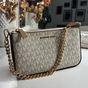 Beige axelväska från Michael Kors - Snygg beige axelväska från Michael Kors med guldfärgad kedja och dragkedja. Väskan har diskret mönster med MK-logga, bruna skinn-detaljer och flera fack inuti för kort och smågrejer. Väskan är i modellen ”Jet set chain pouchette” i färgen ”Vanilla”. Köpt för nypris 1695kr.