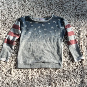 Grå sweatshirt med amerikanska flaggan från Lily White - Cool grå sweatshirt från Lily White i storlek S.  Framsidan har ett blått parti med vita stjärnor och ärmarna har breda röda och vita ränder (USA flaggan / amerikanska flaggan) .  Mjuk insida och rund hals.  Perfekt för dig som gillar statement-plagg och vill sticka ut lite extra. Storlek S/M