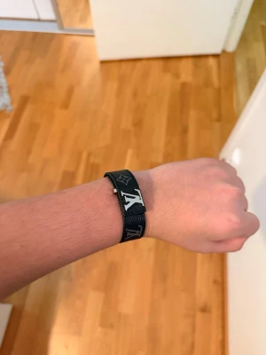 Svart monogram-armband – designer-inspirerat, justerbart, nytt - Stilrent svart armband i monogram-design, inspirerat av klassisk lyxstil. Minimalistisk look med metallspänne och justerbar längd – funkar både till vardag och fest.  🔹 Nytt & oanvänt 🔹 Justerbar storlek 🔹 Levereras i plastpåse (se bilder) 🔹 Lätt och bekvämt att bära