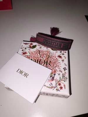 Christian Dior armband - Säljer ett exklusivt västband från Christian Dior i bordeaux och svart med tydlig logotyp. Bandet har snygga fransar i ändarna och är tillverkat i ett slitstarkt syntetmaterial. Perfekt för att ge din väst en lyxig touch. Nypris ca 4000