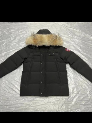 Svart dunjacka från Canada Goose - Svart dunjacka från Canada Goose med avtagbar huva och beige pälsdetalj. Jackan har flera fickor framtill, ribbade muddar och klassisk logga på ärmen. Perfekt för kalla vinterdagar och har en snygg, stilren look.