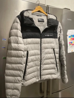 Grå och svart dunjacka Sail Racing - Säljer en dunjacka från Sail Racing i grått och svart med 800 fill power. Jackan har två sidofickor med dragkedja, hög krage och snyggt broderad logga på bröstet. Perfekt för dig som vill ha en varm och lätt jacka med sportig vibe.