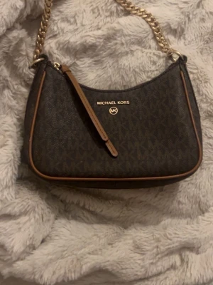 Brun axelväska från Michael Kors - Snygg brun axelväska från Michael Kors med guldfärgad kedja och klassiskt MK-mönster. Väskan har detaljer i skinn och dragkedja upptill. Perfekt storlek för det viktigaste och ger en lyxig touch till din outfit.