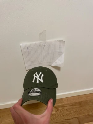 Olivgrön NY keps - Säljer en olivgrön New Era 9FORTY keps med klassisk NY-logga i vitt framtill. Kepsen har böjd skärm och justerbar rem baktill för perfekt passform. Materialet är slitstark bomull och insidan har märkning med 9FORTY och Genuine Merchandise.