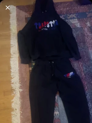 Svarta mjukisbyxor från Trapatar - Säljer ett par svarta mjukisbyxor från Trapstar med broderad logga i rött, vitt och blått på ena benet. Byxorna har resår i midjan med snörning och är tillverkade i mjukt bomullsmaterial. Perfekta för chill dagar eller streetwear-stil.