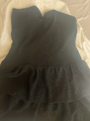 Svart glittrig axelbandslös klänning Missay - Snygg svart axelbandslös klänning från Missay med glittrigt tyg och volangdetaljer nedtill. Kort modell som ger en cool och festlig vibe. Materialet har en skimrande yta som syns tydligt i ljuset. Perfekt för dig som vill sticka ut.