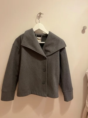 Grå kappa från Gina Tricot - Snygg grå kappa från Gina Tricot med bred krage och tre knappar framtill. Jackan har en rak och loose passform med långa ärmar och är tillverkad i ett mjukt ullmaterial. Perfekt för höst och vinter när du vill ha en stilren look.