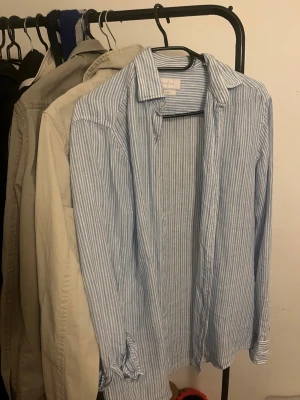 Randig linneskjorta från Massimo Dutti - Fräsch skjorta från Massimo Dutti i 100% linne med ljusblå och vita ränder. Klassisk krage, långärmad och med knappar framtill. Perfekt för dig som gillar en chill och stilren look under varmare dagar.