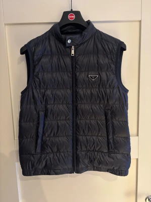 Blå dunväst från Prada - Snygg marin blå dunväst från Prada med glansig design och diskret logga på bröstet. Västen har dragkedja framtill och två fickor. Materialet är polyester med fyllning av vitt gåsdun (90% dun). Perfekt för lager-på-lager under kyliga dagar. Storlek IT 48 vilket motsvarar M. Köpt på Vestaire.   Dragkedjan är lite skadad längst ner, men fungerar fortfarande bra och går att stänga utan problem.  Skickar inom 24h  Priset är inte hugget i sten