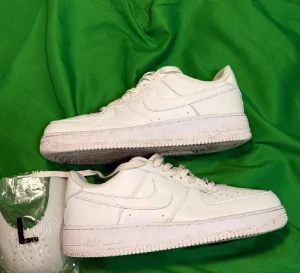 Nike Air Force 1 vita sneakers - Säljer ett par klassiska Nike Air Force 1 sneakers i helvitt skinn med rund tå och platt sula. Skorna har snörning och ikonisk Nike-logga på sidan och hälen. Perfekt för dig som gillar stilrena och tidlösa sneakers. Storlek US 9.5/EU 43.