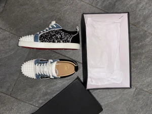Christian Louboutin sneakers med nitar - Säljer ett par unika Christian Louboutin sneakers med vit och blå ovandel, svarta paneler med glittrigt mönster och vita nitar på tån. Skorna har snörning, rund tå och den klassiska röda sulan. Materialmix av skinn och textil, riktigt statement för dig som gillar att sticka ut.