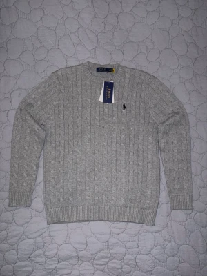 Grå kabelstickad tröja Polo Ralph Lauren - Snygg grå kabelstickad tröja från Polo Ralph Lauren med klassisk rund hals och broderad logga på bröstet. Tröjan har ribbade muddar vid ärmslut och nederkant, och är långärmad. Perfekt för dig som gillar stilrena och tidlösa plagg. Fick den som present men den är för liten för mig så jag har inget kvitto men den är autentisk.