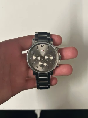 Silverfärgad Tommy Hilfiger klocka - Säljer en stilren klocka från Tommy Hilfiger i silverfärgat rostfritt stål. Klockan har en rund urtavla med tre mindre visartavlor, tydliga siffror och Hilfiger-loggan. Länken är bred och robust, perfekt för dig som gillar klassisk design.