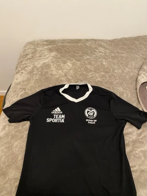 Svart fotbollströja Motala AIF Adidas - Säljer en svart fotbollströja från Adidas med Motala AIF tryck och TEAM SPORTIA logga på bröstet. Tröjan har vit krage och nummer 7 på ryggen. Perfekt för dig som gillar sportiga och klassiska lagtröjor.
