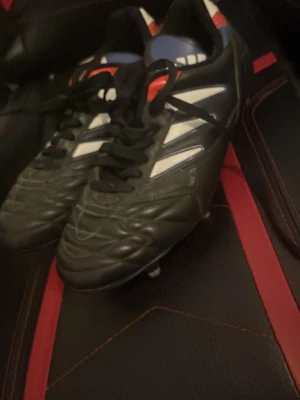 Addidas predator absoulute från 2000 talet med nyckel - Säljer ett par svarta Adidas fotbollsskor med vita och röda detaljer. Skorna har snörning och metall-dobbar för extra grepp på planen. Ovandelen är tillverkad i skinn och har en klassisk rund tå. Perfekt för dig som vill ha stabilitet och stil på fotbollsplanen.