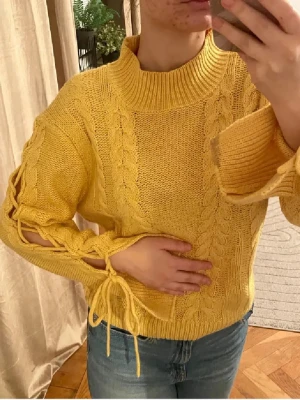 Gul stickad tröja med snörning - Säljer en gul stickad tröja med grova flätstickade detaljer och snörning längs ärmarna. Tröjan har hög krage och långa ärmar med öppna partier och knyt längst ner. Perfekt för dig som gillar färg och unika detaljer.