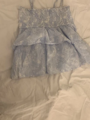 Ljusblå linnetopp med volanger - Säljer en ljusblå linnetopp med smala axelband och volangdetaljer. Toppen har ett paisleyinspirerat mönster och smock upptill för en skön passform. Perfekt för varma dagar och sommarhäng.