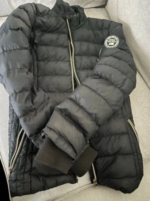 Svart pufferjacka från Vinson Polo Club - Snygg svart pufferjacka för flickor från Vinson Polo Club med quiltad design och ribbade muddar. Jackan har silverfärgade dragkedjor och ett stort märke på ärmen. Perfekt för kalla dagar och ger en cool streetstil. Jackans huva saknas!!!!!!  Tveka inte att höra av dig om du har några funderingar. 