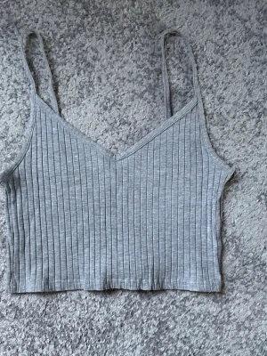 Grå ribbad linnetopp med smala axelband - Säljer en ljusgrå croppad linnetopp med smala axelband och ribbad struktur. Toppen har v-ringad hals och är perfekt för varma dagar. Enkel och stilren design som är lätt att matcha med olika outfits.