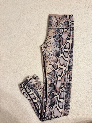 Ormmönstrade leggings från Aim'n - Säljer ett par snygga leggings från Aim'n med ormmönster i beige, brun och svart. De har hög midja och är gjorda i ett stretchigt syntetmaterial som sitter skönt och följer kroppen. Perfekta för träning eller chill. Mönstret är riktigt coolt och sticker ut.
