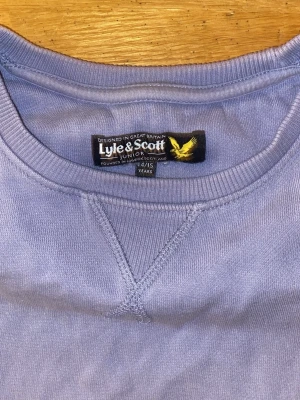 Ljuslila sweatshirt från Lyle & Scott - Säljer en ljuslila sweatshirt från Lyle & Scott med klassisk rund hals och ribbade muddar. Tröjan har ett broderat gult logomärke på bröstet och är tillverkad i mjuk bomull. Perfekt för dig som gillar stilrena och bekväma plagg.