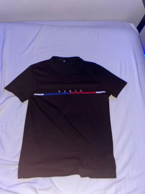 Svart t-shirt med Paris-tryck - Svart t-shirt med rund hals och korta ärmar. Framtill finns ett tryck med texten 'PARIS' och en horisontell linje i blått, vitt och rött. Materialet känns som mjuk bomull, perfekt för en chill och stilren look.
