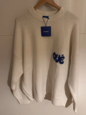 Vit stickad tröja från Qué Sera Sera - Vit stickad tröja med blått broderat Qué-tryck på bröstet. Tröjan har rund hals, ribbade muddar och en loose passform. Mjuk och skön känsla, perfekt för kyliga dagar. Enkel design med en liten twist tack vare det blå trycket.