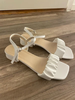 Vita sandaler med låg klack, strl 38-41 - Snyggaste vita klackar med fyrkantig tå och bekväm blockklack. Har en mjuk rynkad rem framtill och justerbart spänne runt ankeln så de sitter stabilt. Perfekta till sommarfester, student, bröllop eller när du vill va lite extra clean ✨De är i storlek 41 men skulle säga att de passar 38-39 och de har aldrig kommit till användning så de är i mycket bra skick😌
