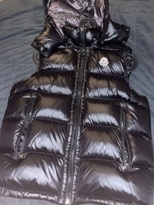 Svart dunväst från Moncler - Säljer en svart, glansig dunväst från Moncler med huva och två praktiska fickor med dragkedja. Västen har quiltad design och Moncler-logga på bröstet. Perfekt för kyliga dagar och har en snygg, modern look med dragkedja framtill. inget fel på den.