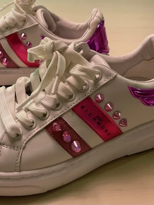John Richmond sneakers med rosa nitar - Säljer ett par vita sneakers från John Richmond med glansiga rosa detaljer och nitar på sidorna. Skorna har tjock vit sula, snörning och metallicrosa hälparti med JR-logga. Materialet är skinn och syntet, och designen är riktigt eye-catching med coola spikes och glitter.läder, äkta.