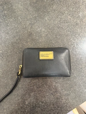 Svart plånbok från Marc Jacobs - Snygg svart plånbok från Marc Jacobs i äkta skinn med guldfärgade metalldetaljer och dragkedja runtom. Plånboken har en praktisk handledsrem och en metallbricka med logga på framsidan. Perfekt storlek för att ha med sig det viktigaste. Köptes för ungefär 3000 kronor för ett tag sedan men är fortfarande i bra skick 